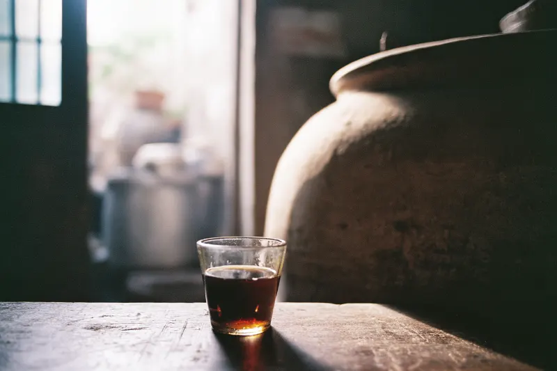 A glass of amber sealed-vat rice wine with a clay vat behind, Jiangsu